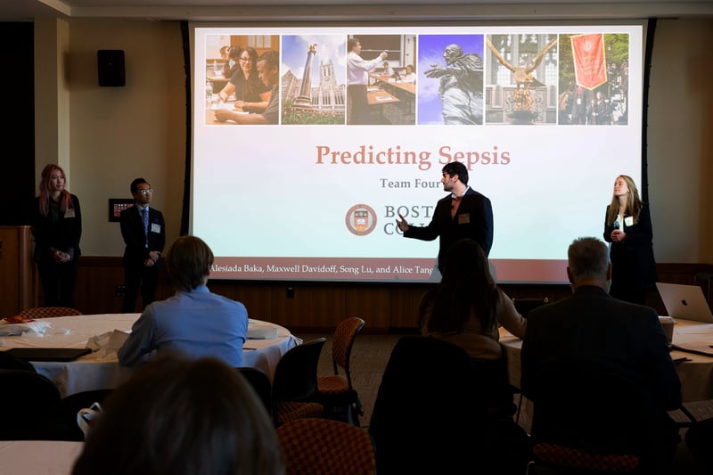 A team from Boston College presents on "Predicting Sepsis" in a formal setting, with a projected slide and an audience watching their discussion.