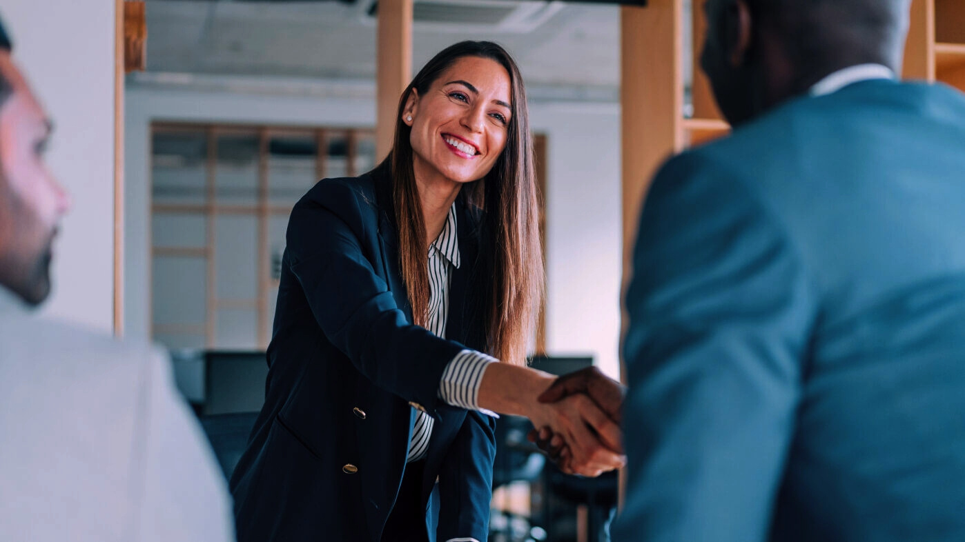 A professional woman in a business suit smiles while shaking hands with a colleague in a modern office setting, symbolizing successful collaboration, networking, or a business agreement.